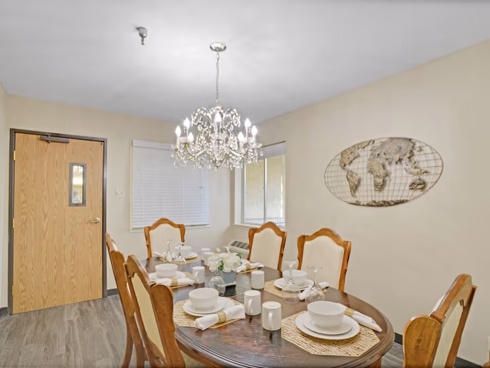 A dining room with a wooden oval table set for six people. The table is arranged with white bowls, plates, napkins, glasses, and small decorative items. Six wooden chairs with cream-colored cushions surround the table. A crystal chandelier hangs above the table, and there is a wooden door and two windows with blinds on the far wall. A metal wall decoration in the shape of a world map is mounted on the right wall.