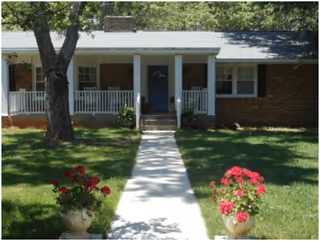 Single-story brick house with a white front porch and blue door, surrounded by green grass and trees. Two flower pots with red flowers are placed symmetrically on either side of a concrete walkway leading to the porch.