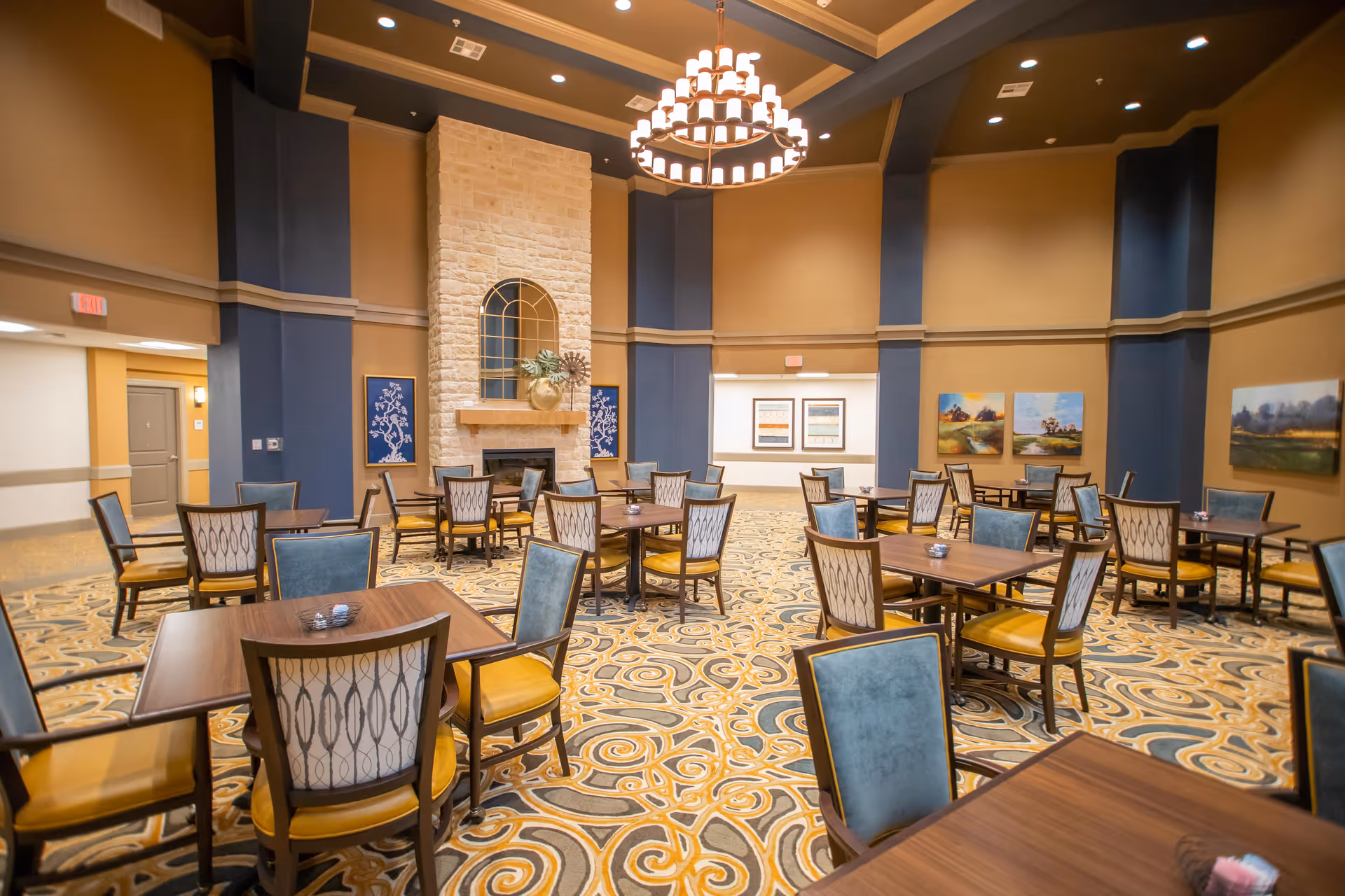 A spacious dining room with multiple wooden tables and chairs arranged neatly on a patterned carpet. The room features a tall stone fireplace with decorative items on the mantel, warm beige and blue walls, several framed artworks, and a large chandelier hanging from the ceiling.