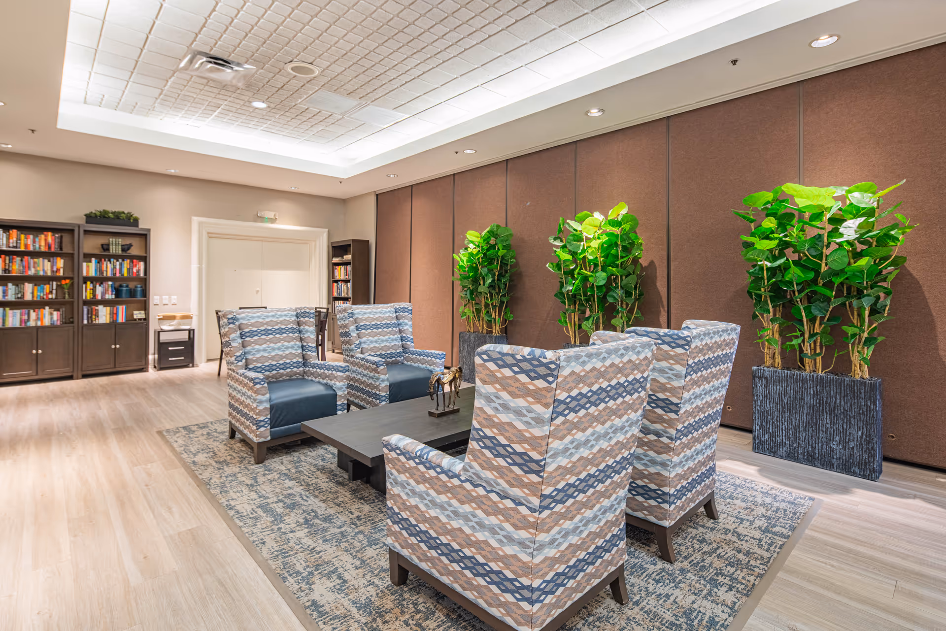 A bright and spacious lounge area with six patterned armchairs arranged around a low rectangular coffee table on a patterned rug. Behind the chairs are three large potted green plants against a brown wall. To the left, there are bookshelves filled with books and decorative items. The ceiling has recessed lighting and a textured design.