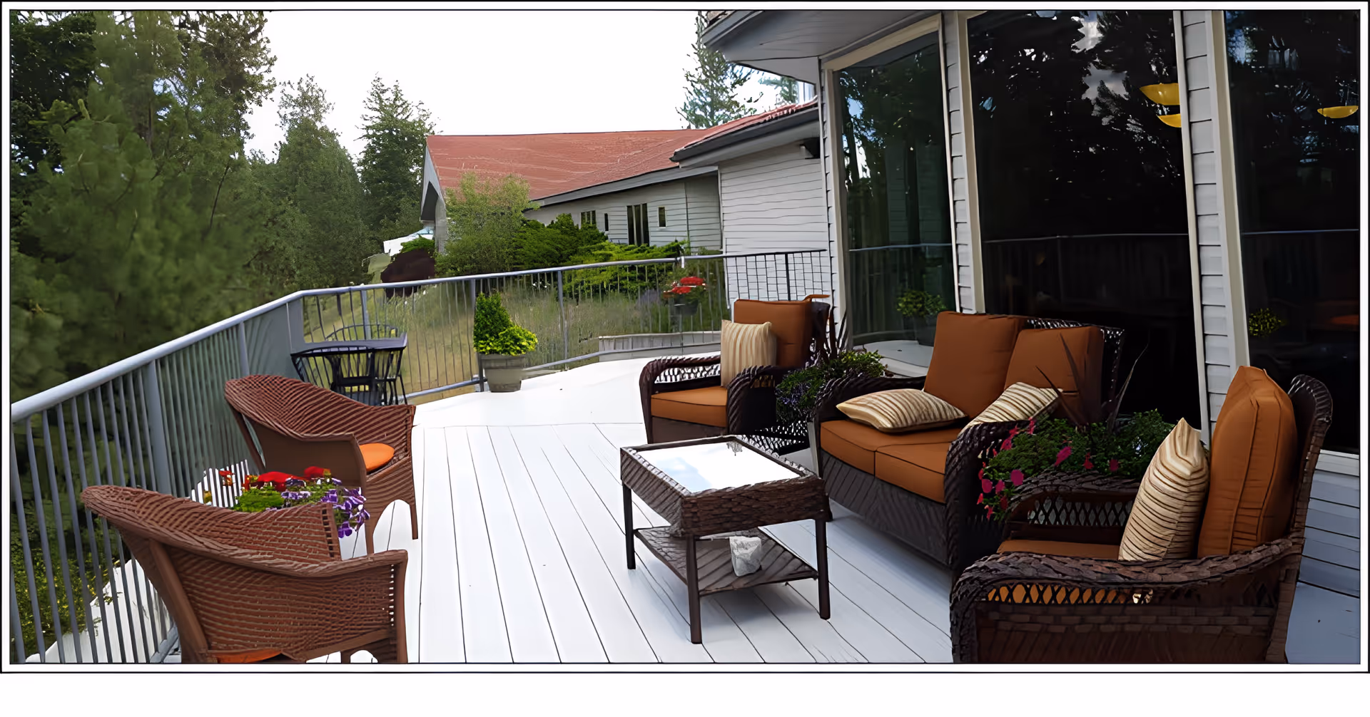 Outdoor patio area with brown wicker furniture including chairs and a loveseat with orange cushions and striped pillows. A glass-top wicker coffee table is in the center. The patio has white flooring and is surrounded by a metal railing. There are potted plants with flowers on the patio and trees and greenery in the background.