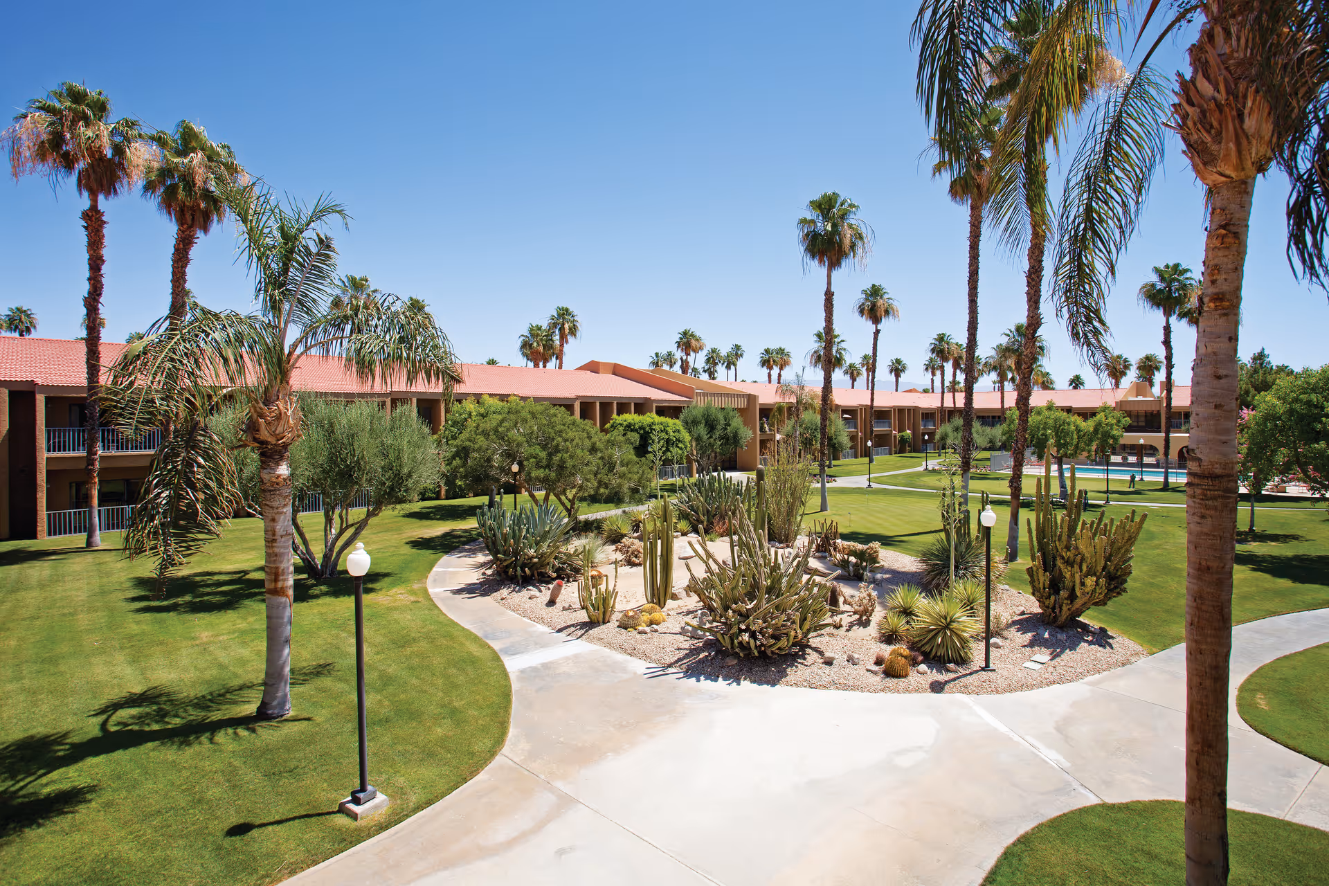 Outdoor view of The Carlotta, Ivy Signature Living facility featuring a landscaped garden with palm trees, cacti, and green lawns under a clear blue sky. The building with red-tiled roofs surrounds the garden area, and paved walkways curve through the lawn.