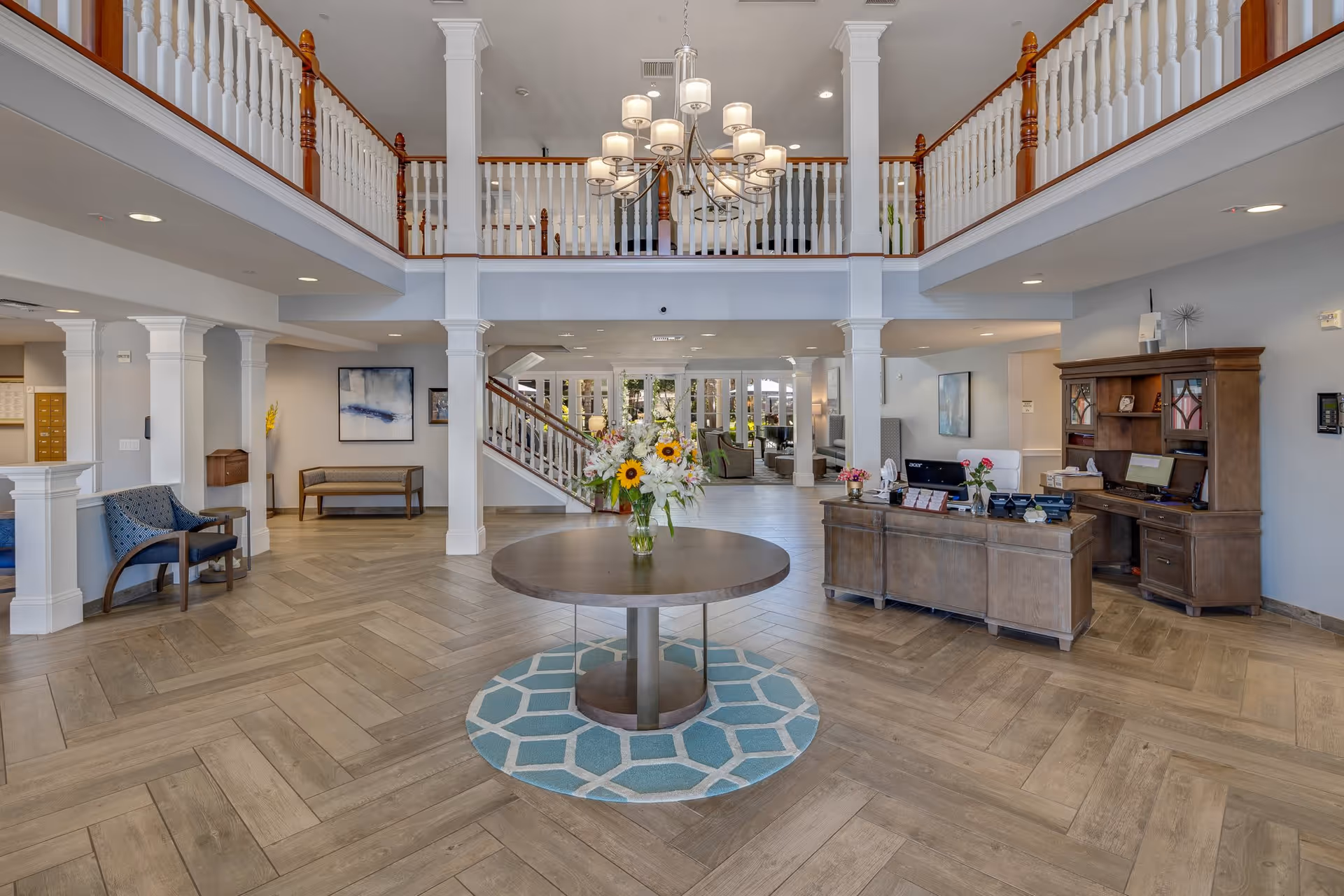 Spacious senior living facility lobby with a round table holding a vase of flowers in the center on a blue patterned rug. To the right is a wooden reception desk with office equipment and flowers. The area features light wood flooring, white columns, a staircase with wooden railings, and a chandelier hanging from the ceiling. Seating areas and artwork are visible in the background.