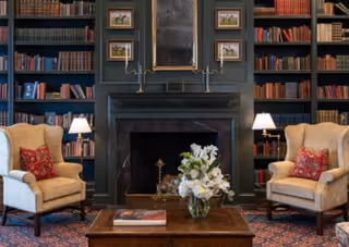 A cozy living room with two beige armchairs featuring red patterned cushions, positioned on either side of a dark fireplace. The fireplace is surrounded by built-in bookshelves filled with books and framed pictures. A wooden coffee table with a vase of white flowers and a book sits in front of the fireplace. Two table lamps with white shades provide warm lighting.