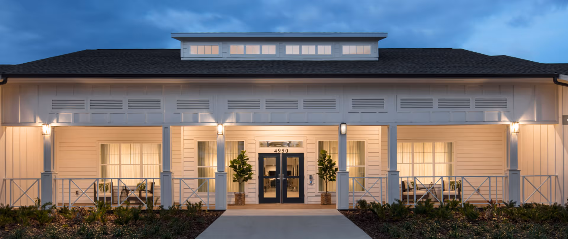 Front entrance of a white single-story building with a covered porch, double glass doors, potted plants, and warm exterior lighting.