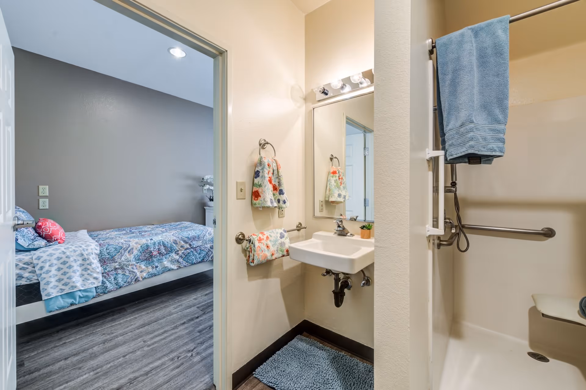 View of a bathroom with a small white sink, a mirror above it, and colorful floral hand towels hanging on the wall. To the right is a shower area with a blue towel hanging on the grab bar and a fold-down seat inside the shower. Through an open door, a bedroom with a bed covered in a blue and white patterned bedspread is visible.