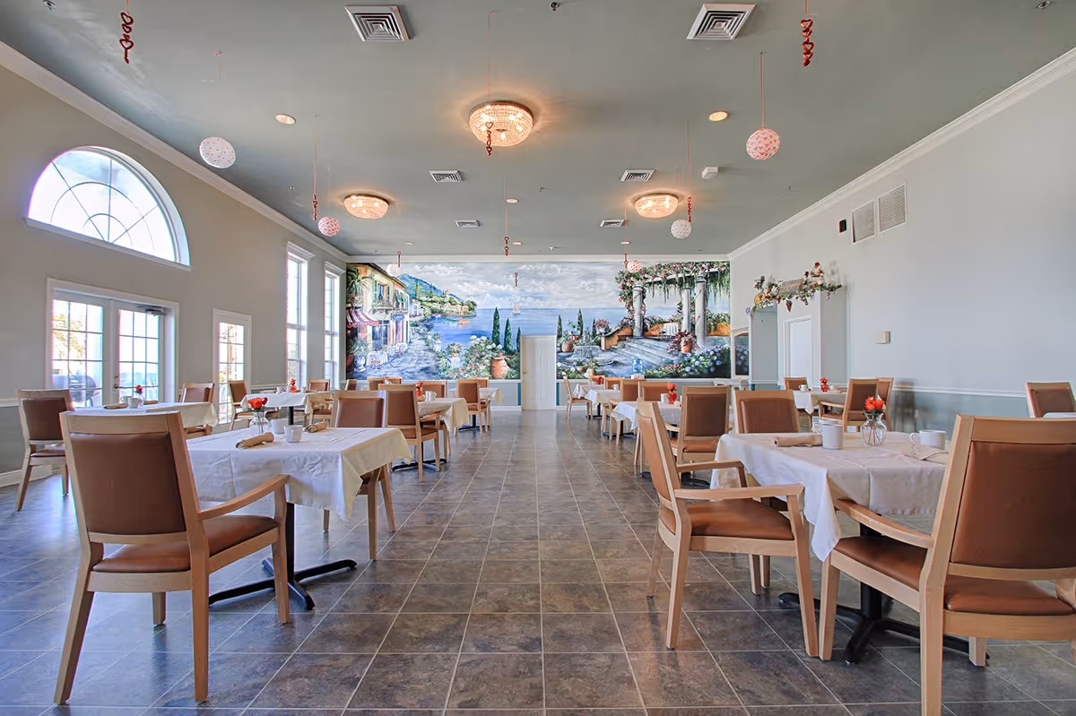 A spacious dining room with multiple tables covered in white tablecloths, each set with cups, napkins, and small flower vases. The room features large windows and a door on the left side, allowing natural light to enter. The far wall has a large mural depicting a scenic coastal landscape with greenery, buildings, and a blue sky. The ceiling has several round light fixtures and hanging decorative ornaments.
