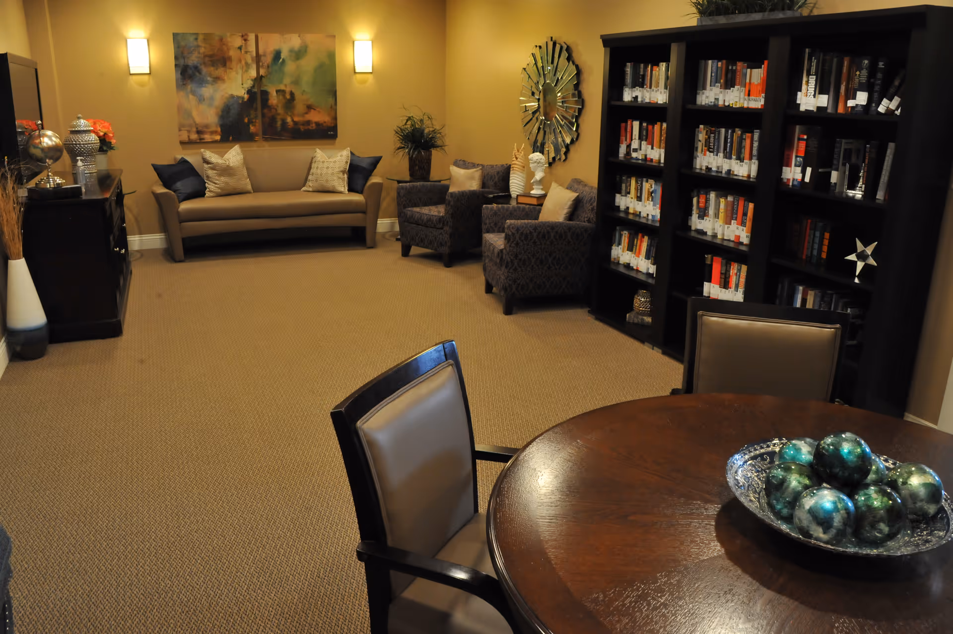 A cozy living room area with a beige sofa, two patterned armchairs, a round wooden table with chairs, and a large bookshelf filled with books. The walls are painted warm yellow and decorated with a large abstract painting and a decorative mirror. Soft lighting fixtures illuminate the room.