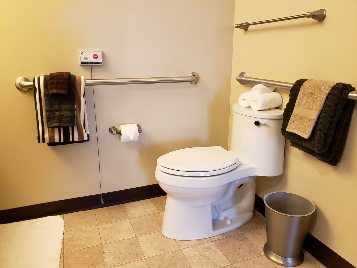 A clean bathroom with a white toilet, beige walls, and tiled floor. There are metal grab bars on the walls with folded towels hanging on them. A small trash can is placed on the floor next to the toilet, and a white bath mat is partially visible on the floor. A toilet paper holder with a roll of toilet paper is mounted on the wall.