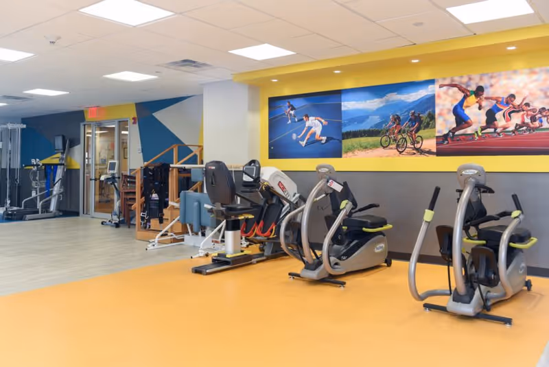 A rehabilitation and fitness room with various exercise machines including stationary bikes and other equipment. The room has a yellow and gray color scheme with a large mural on the wall depicting people playing tennis, cycling, and running. The floor is a combination of light wood and yellow rubberized flooring.