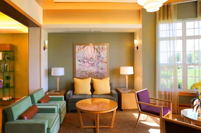 A bright and cozy living room with a gray sofa adorned with two yellow pillows, flanked by two side tables each holding a lamp. In front of the sofa is a round wooden coffee table. To the left are two green armchairs with brown cushions, and to the right is a purple armchair. Large windows with sheer curtains allow natural light to fill the room. A colorful abstract painting hangs on the wall behind the sofa.