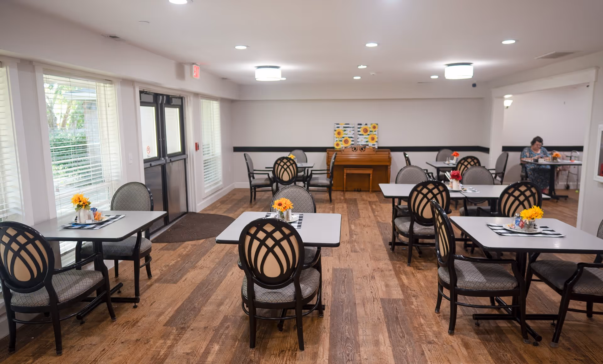 A dining room in a senior living facility with several tables and chairs arranged neatly. Each table has a small flower arrangement. There is a piano against the far wall with sunflower paintings above it. A woman is seated at a table on the right side of the room. Large windows and a double door provide natural light on the left side.