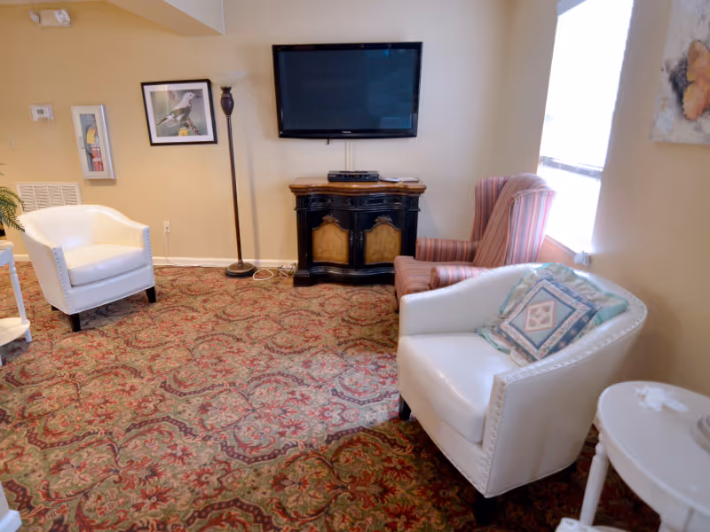 A cozy living room with patterned carpet, two white armchairs, a striped armchair near a window, a small round white side table, a floor lamp, a framed bird picture on the wall, and a flat-screen TV mounted above a dark wooden cabinet.