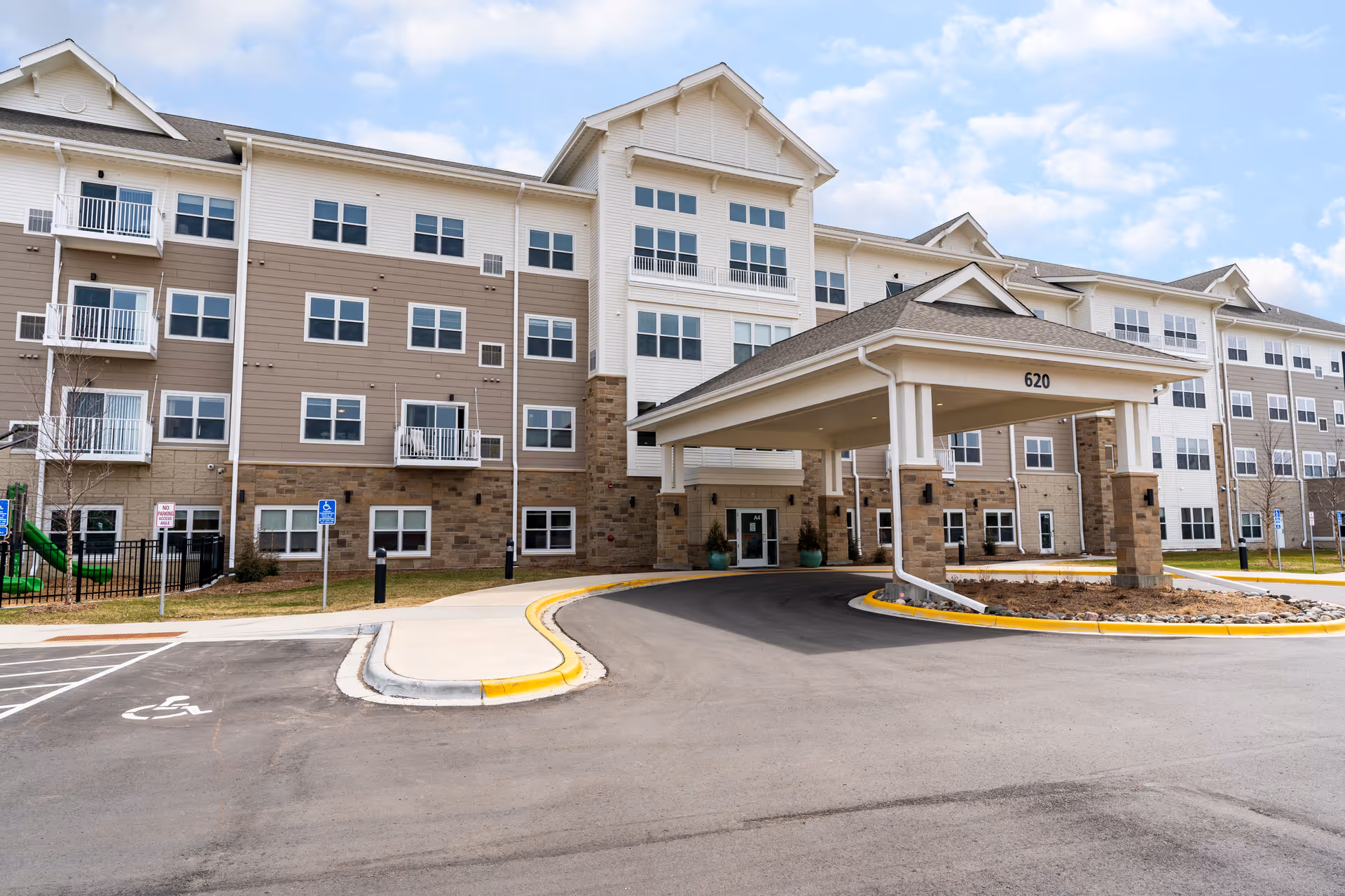 Exterior view of Riley Crossing Senior Living facility showing a large multi-story building with beige and white siding, multiple windows, balconies, and a covered entrance with the number 620 above it. The foreground includes a driveway, parking spaces, and a sidewalk with a wheelchair accessible sign.