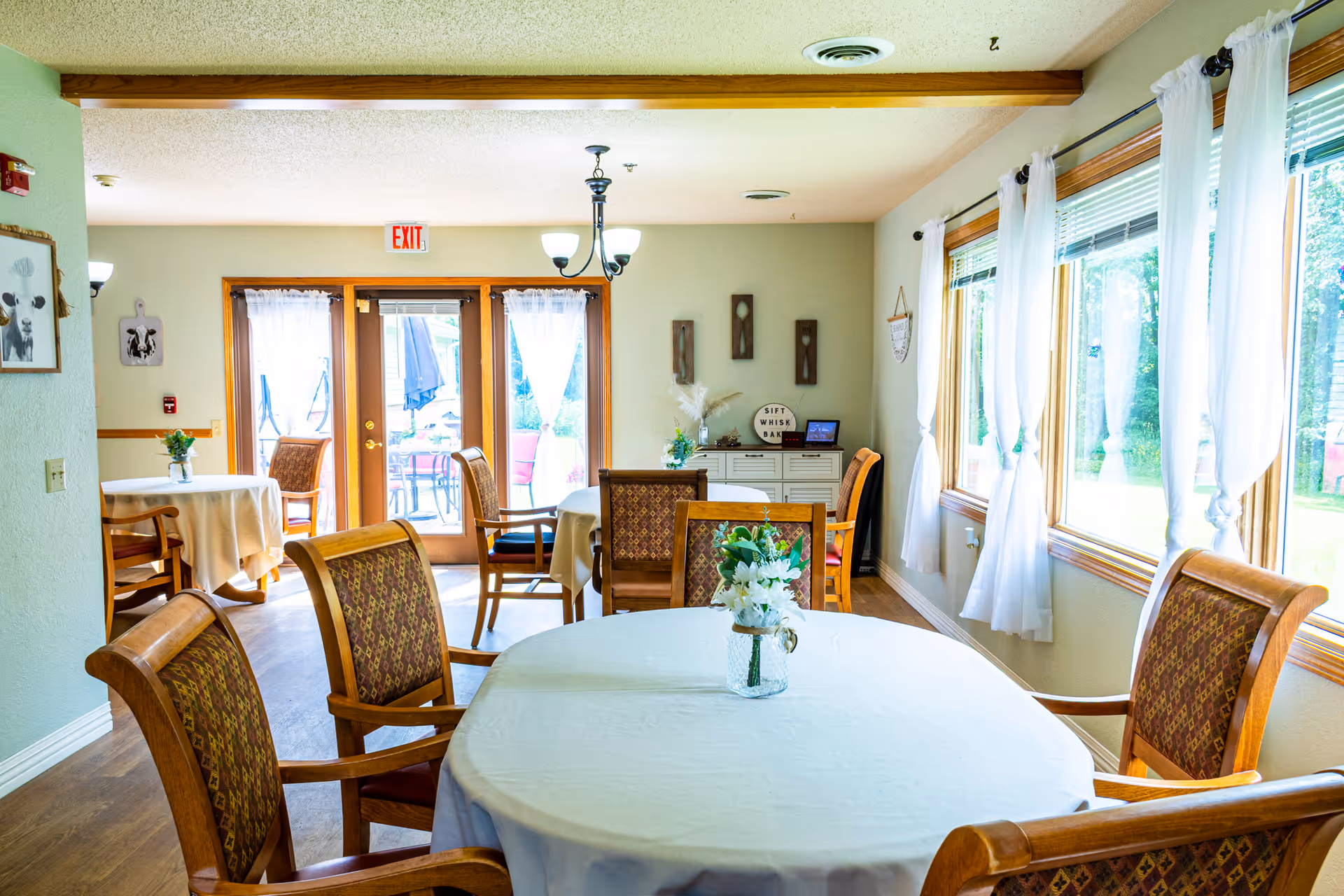 A bright dining room in a senior living facility with round tables covered in white tablecloths, wooden chairs with patterned cushions, and a vase with white flowers on the nearest table. Large windows with white curtains allow natural light to fill the room, and glass doors lead to an outdoor patio area with seating. The walls are light-colored with simple decorations, and a chandelier hangs from the ceiling.