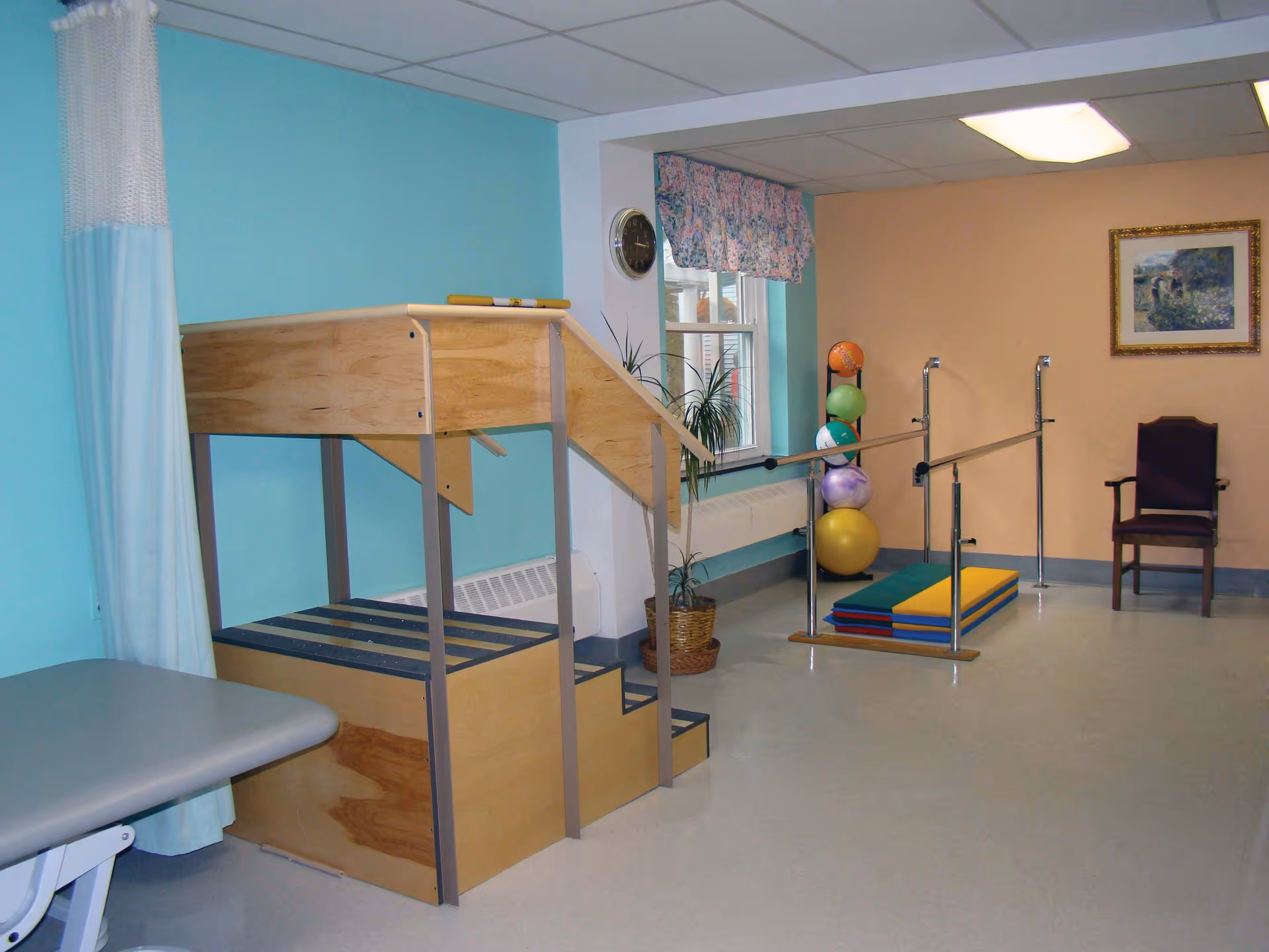 A physical therapy room with wooden stairs and handrails for rehabilitation exercises, colorful exercise balls stacked in a corner, padded mats on the floor, a chair against the wall, a window with floral curtains, and a clock on the wall.