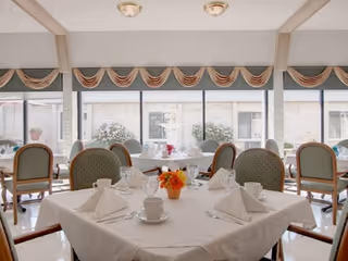 A dining room with a table set for four, featuring white tablecloth, folded white napkins, cups, glasses, and a small orange flower centerpiece. The room has large windows with valance curtains and several chairs around the table.