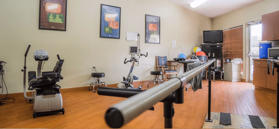 A rehabilitation room with exercise equipment including a stationary bike, parallel bars for walking practice, stools, chairs, and therapy balls. The room has wooden flooring, light-colored walls with framed motivational posters, and a TV on a stand near a window with blinds.