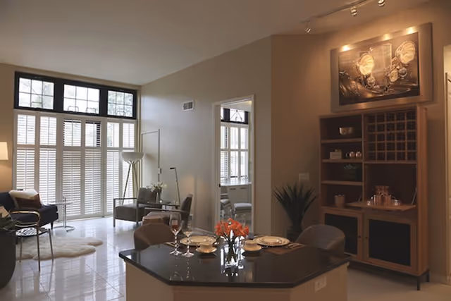 A modern living and dining area with large windows covered by white shutters allowing natural light to fill the room. The space features a black countertop dining table set with plates, bowls, glasses, and a vase of orange flowers. There are comfortable chairs around the table, a wooden shelving unit with wine storage and decorative items, and a cozy seating area with armchairs and a floor lamp near the windows.