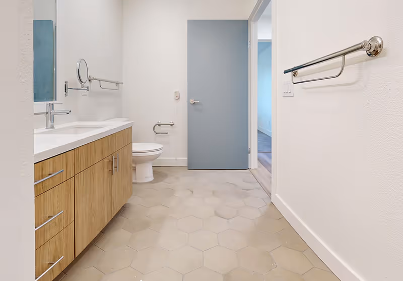 A modern bathroom with a wooden vanity featuring a white countertop and sink on the left, a toilet next to a blue door at the back, hexagonal floor tiles, and metal grab bars on the walls.