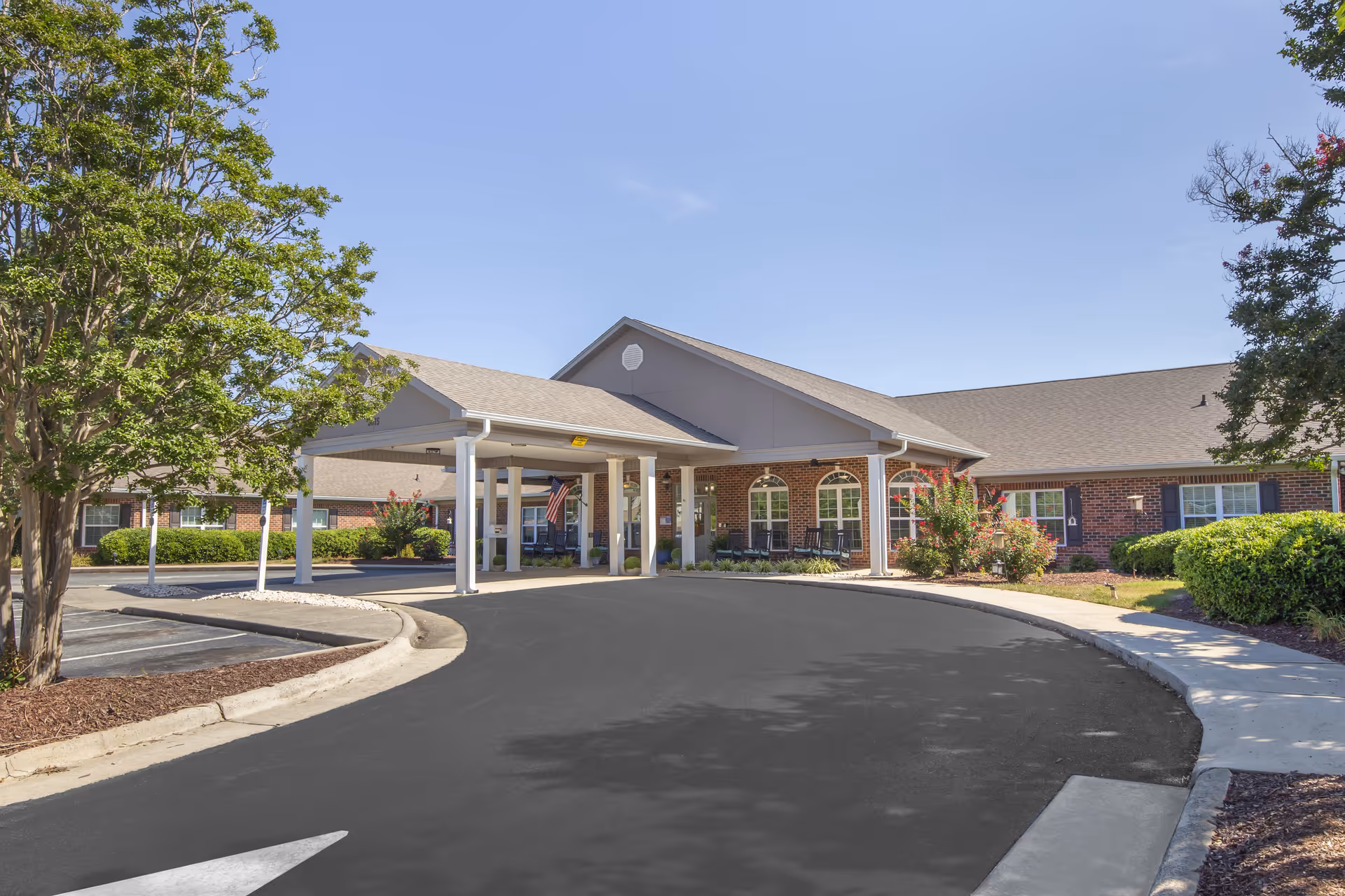 Exterior view of a single-story brick building with a covered entrance driveway, surrounded by landscaped bushes and trees under a clear blue sky.