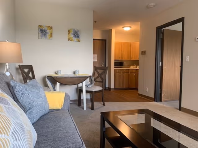 A cozy living room area with a gray couch adorned with patterned cushions and a yellow throw pillow. A small wooden table with two chairs is placed against the wall, which has two floral paintings. In the background, there is a kitchen area with wooden cabinets, a microwave, and a sink. To the right, there is an open doorway leading to another room.