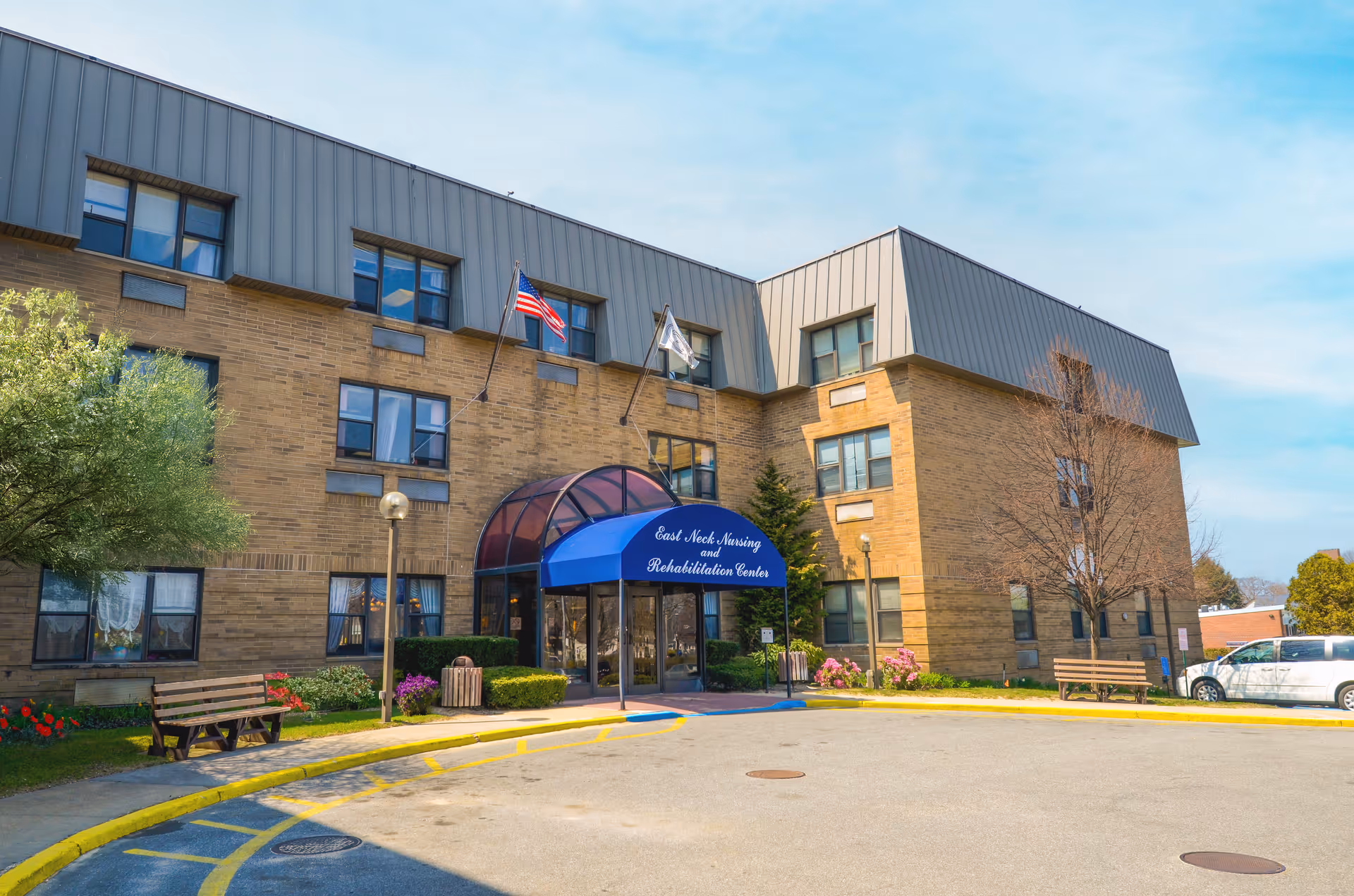 Front entrance of a three-story brick nursing and rehabilitation center with a blue awning, flags, benches, and landscaped beds.