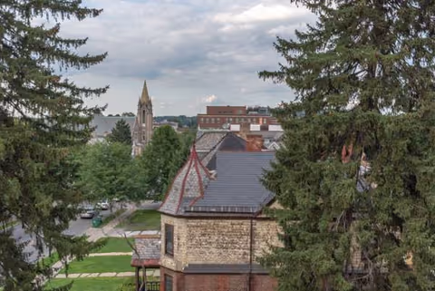 Outdoor view of a brick building with a steep roof, surrounded by trees and distant spires.