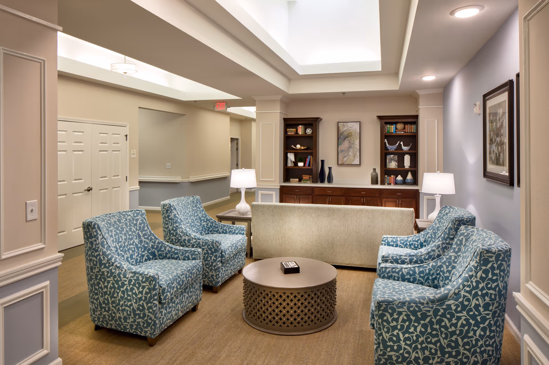 A cozy sitting area in an assisted living facility featuring four blue patterned armchairs arranged around a round coffee table, with a beige sofa facing two wooden bookshelves filled with decorative items and books. The room has soft lighting from table lamps and a skylight above, with framed artwork on the walls and a neutral color palette.