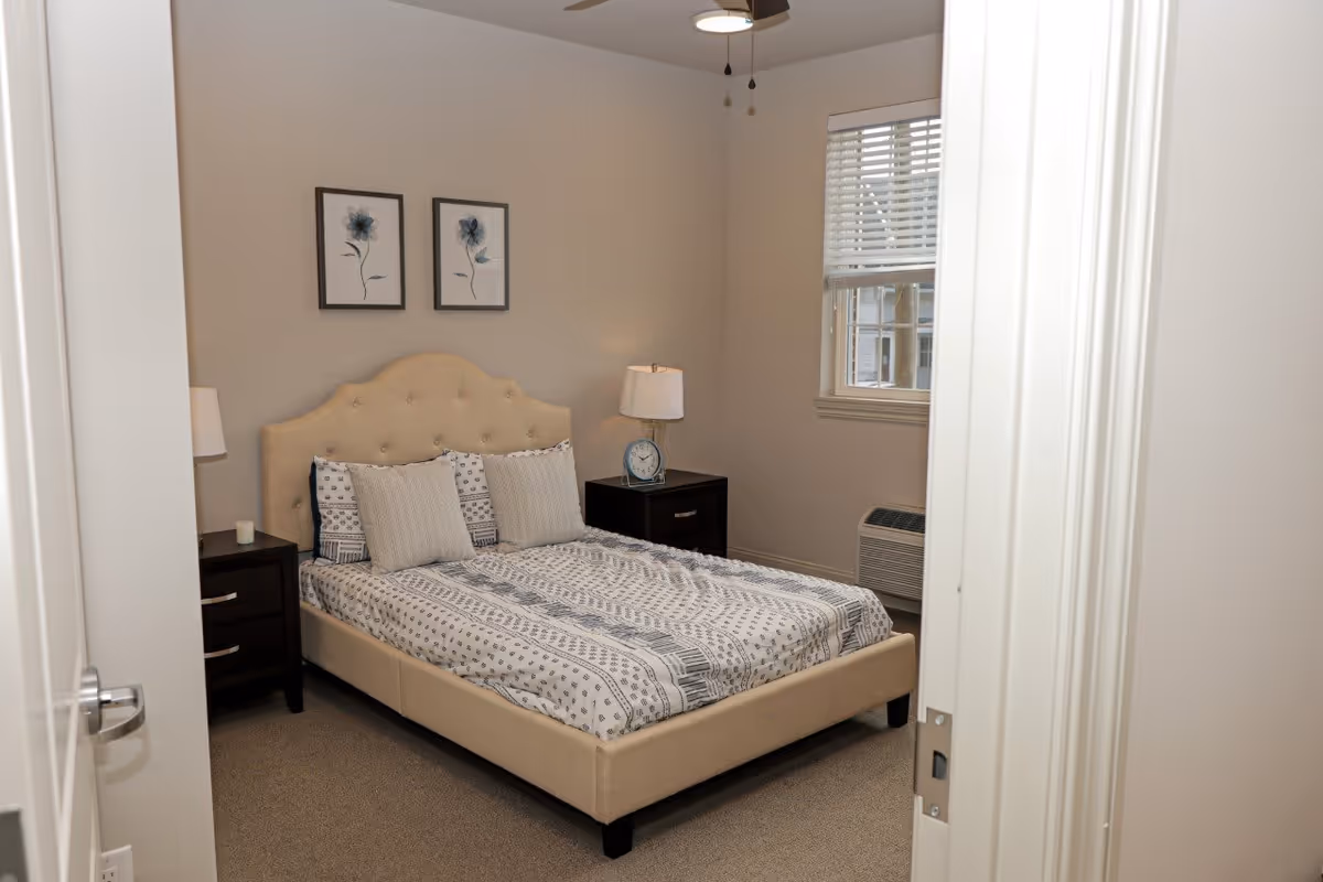 Neatly made bedroom with a beige upholstered bed, patterned bedding, two bedside tables with lamps, and framed floral prints above the headboard.