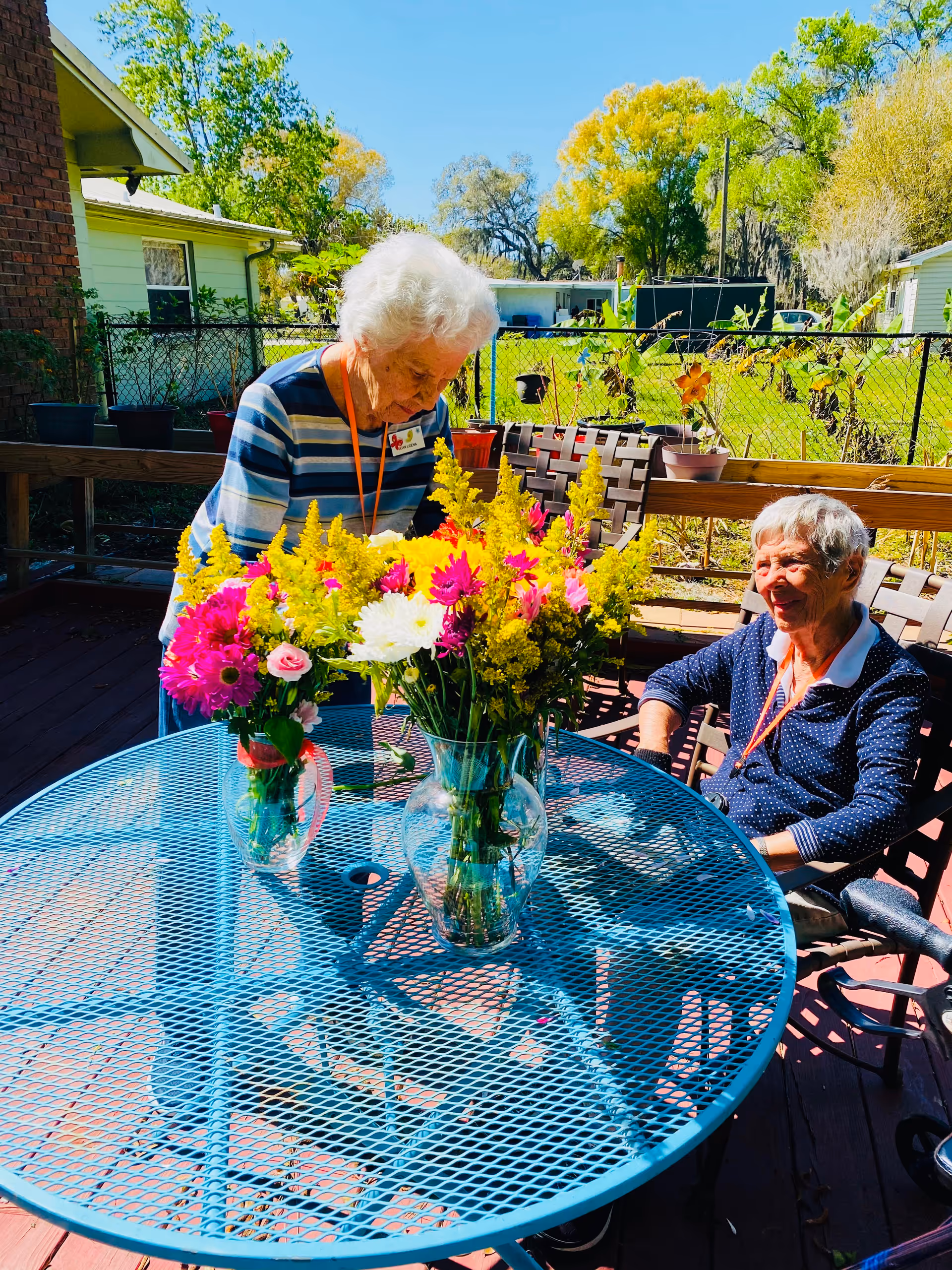 Two elderly women on a sunny outdoor patio with a blue metal table in front of them. One woman is standing and arranging colorful flowers in a vase on the table, while the other woman is sitting and smiling. The background shows a fenced yard with green grass, trees, and neighboring houses under a clear blue sky.