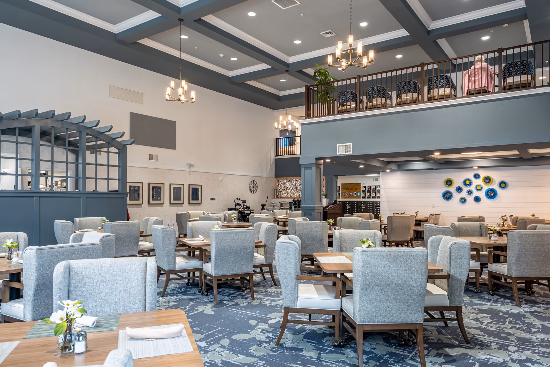 A spacious dining room in a senior living facility with multiple tables and cushioned chairs arranged neatly. The room features a high ceiling with modern chandeliers, a patterned carpet, and a mezzanine level with additional seating. There are decorative wall art pieces and a small plant on the mezzanine railing. The overall color scheme includes shades of blue, gray, and beige.
