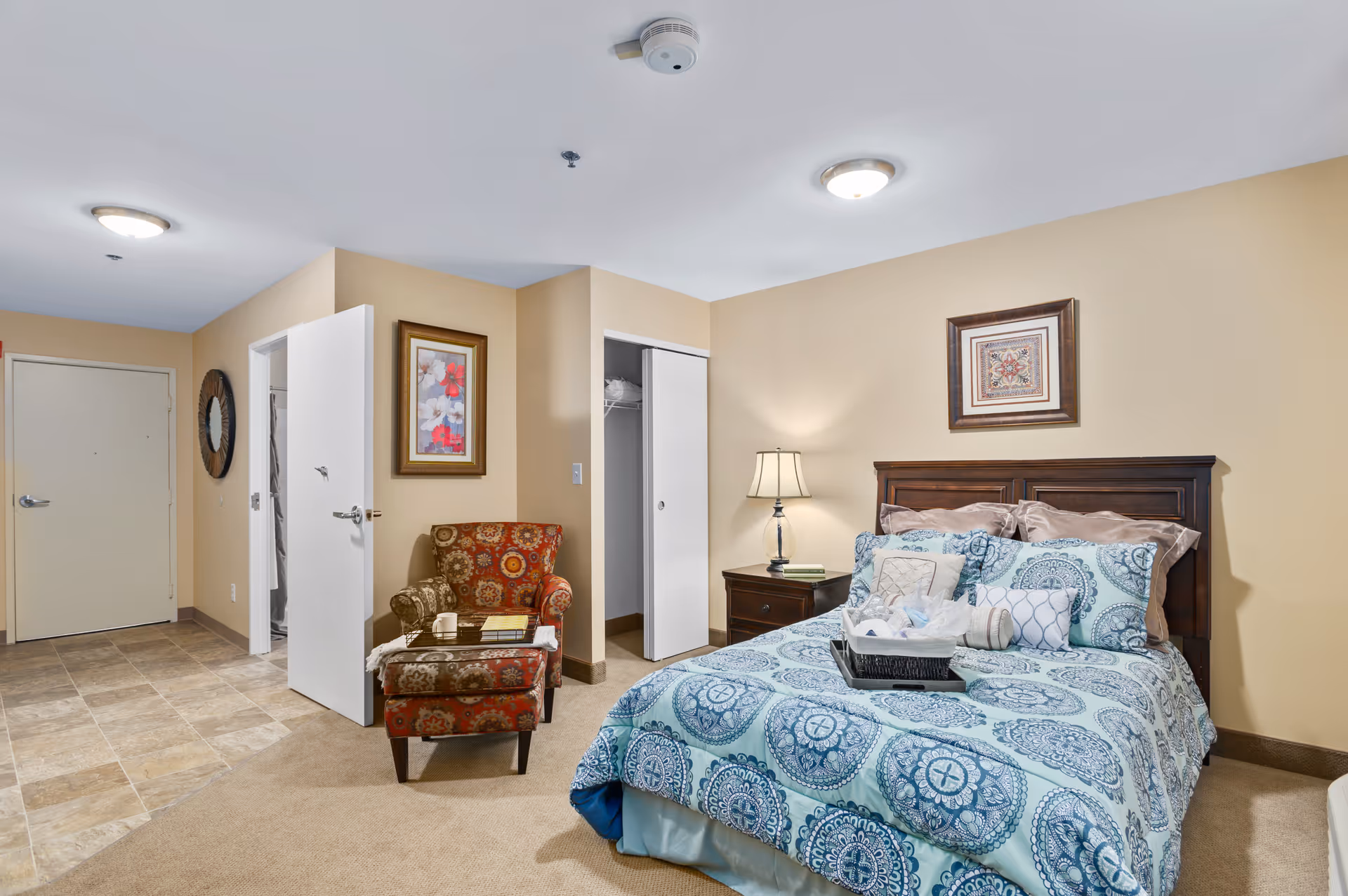 Well-lit furnished bedroom with a patterned bed, nightstand and lamp, an upholstered chair, and open closet and entry door.