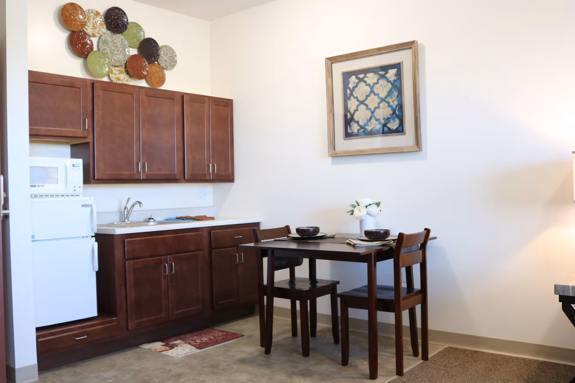 A small kitchen area with dark wooden cabinets, a white mini refrigerator, and a microwave. Next to the kitchen is a dark wooden dining table set for two with bowls, napkins, and a small vase of white flowers. The walls are light-colored with decorative wall art above the cabinets and the dining table.