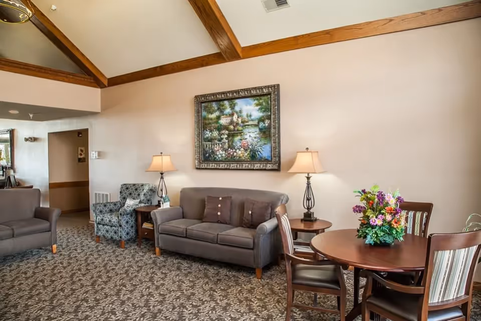 A cozy living room area with a gray sofa, a patterned armchair, two side tables with lamps, a round wooden table with four chairs, and a colorful flower arrangement on the table. The walls are beige with wooden beams on the ceiling and a framed painting of a garden and water scene hanging above the sofa.
