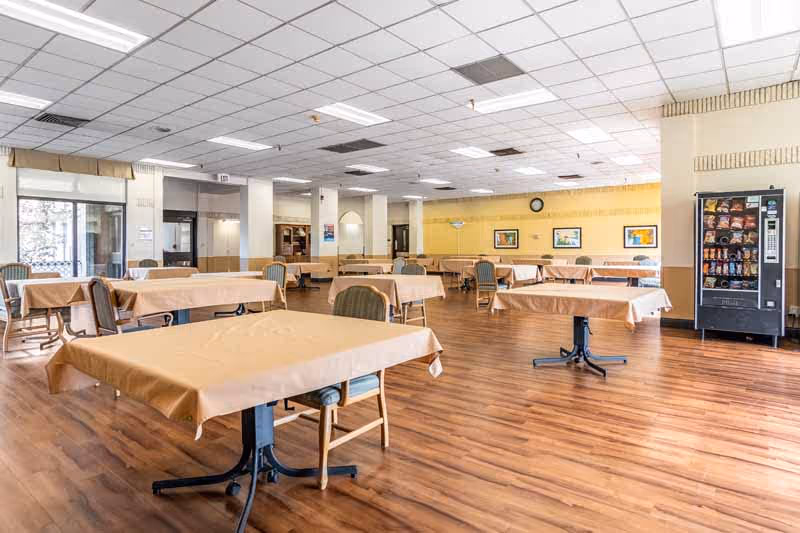 A spacious dining room with multiple tables covered in beige tablecloths and surrounded by chairs. The room has wood flooring, a drop ceiling with fluorescent lights, and a vending machine on the right side. There are framed pictures and a clock on the far yellow wall.