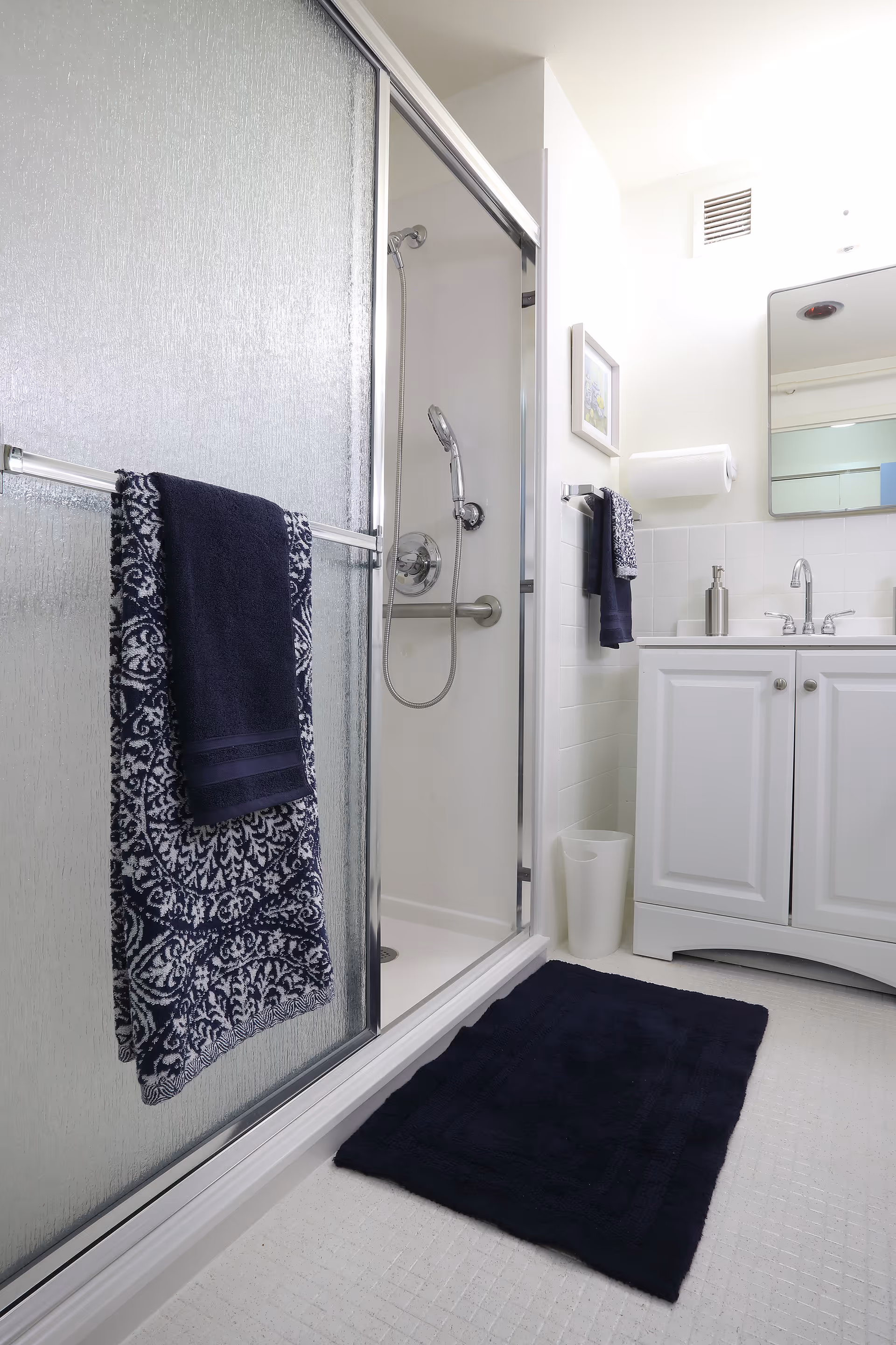 A clean and bright bathroom featuring a glass-enclosed shower with a handheld showerhead, a white vanity with a sink and faucet, a mirror above the sink, a towel rack with two dark blue patterned towels, a matching dark blue bath mat on the floor, a wall-mounted paper towel holder, and a small trash bin beside the vanity.