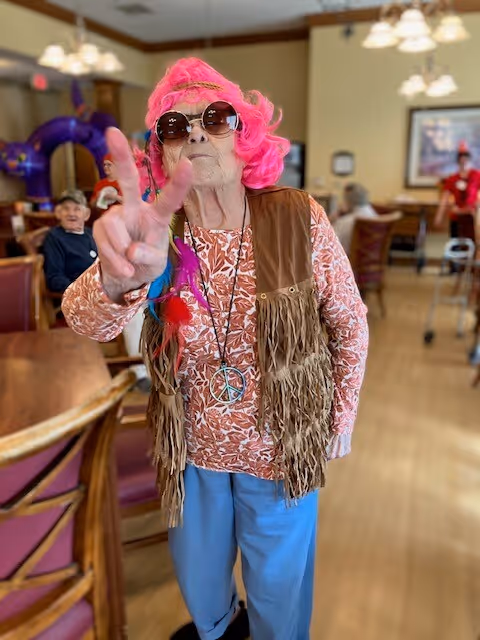 An elderly woman wearing a bright pink wig, large round sunglasses, a fringed brown vest, and a peace sign necklace poses with a peace hand gesture in a senior living facility dining room. Other seniors and dining tables are visible in the background.