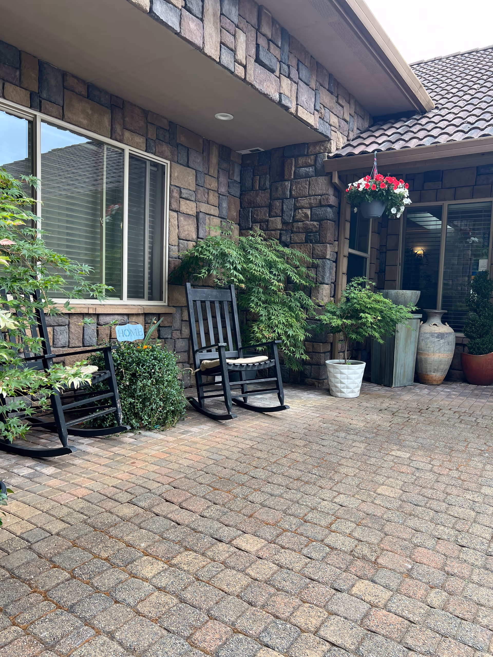 Outdoor patio area with stone walls and tiled roof, featuring two black wooden rocking chairs, several potted plants including a hanging basket with red and white flowers, and a small sign that says 'HOME'.
