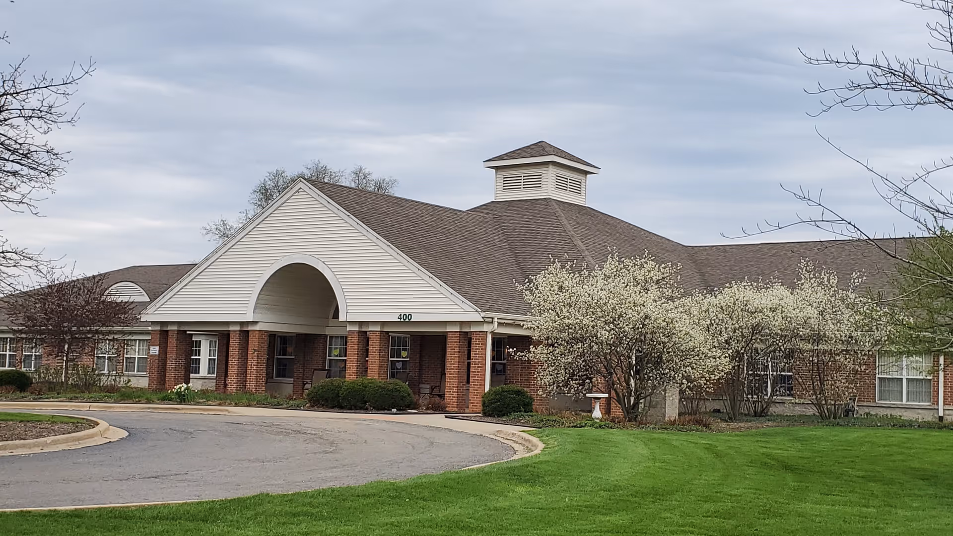 Exterior view of a single-story brick building with a large covered entrance and a circular driveway. The building has a peaked roof with a small cupola on top. There are blooming white flowering trees and well-maintained green grass in front of the building under a cloudy sky.