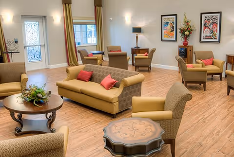 A spacious and well-lit living room area with multiple beige and brown upholstered chairs and sofas, some with red accent pillows. The room has wooden flooring, a round wooden coffee table with a plant centerpiece, and a decorative side table. The walls are adorned with framed artwork and a floral arrangement on a small cabinet. There is a door with decorative glass panels and windows with curtains allowing natural light to enter.