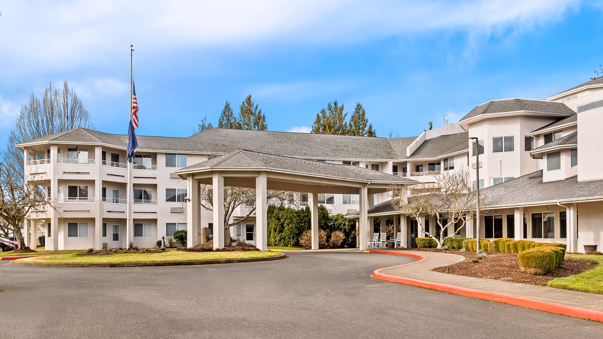 Exterior view of Holiday Sheldon Oaks senior living facility showing a large multi-story building with a covered entrance driveway, manicured bushes, trees, and an American flag at half-mast against a blue sky.
