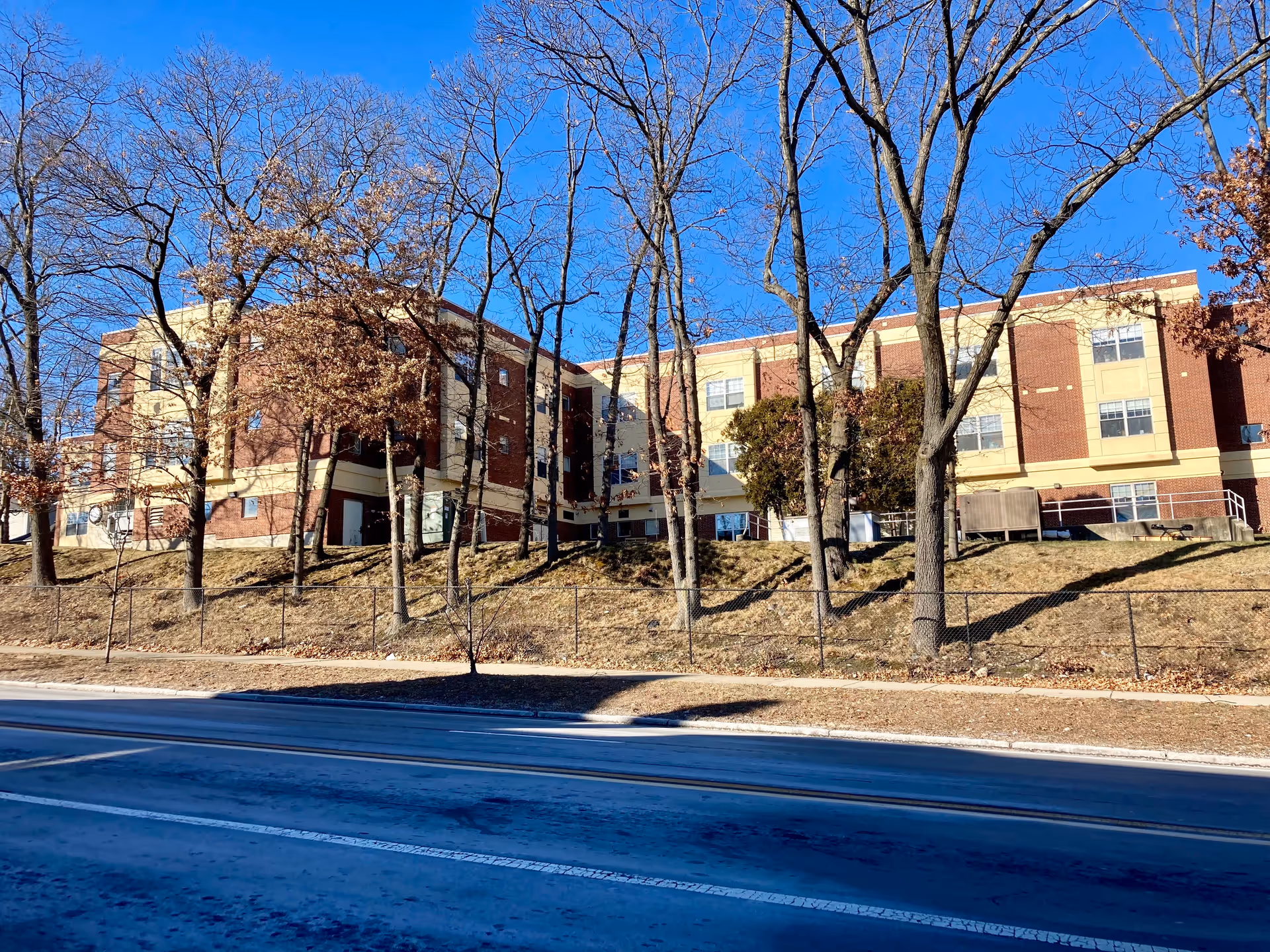 Exterior view of a three-story brick and beige building behind leafless trees and a chain-link fence on a clear sunny day with a bright blue sky.