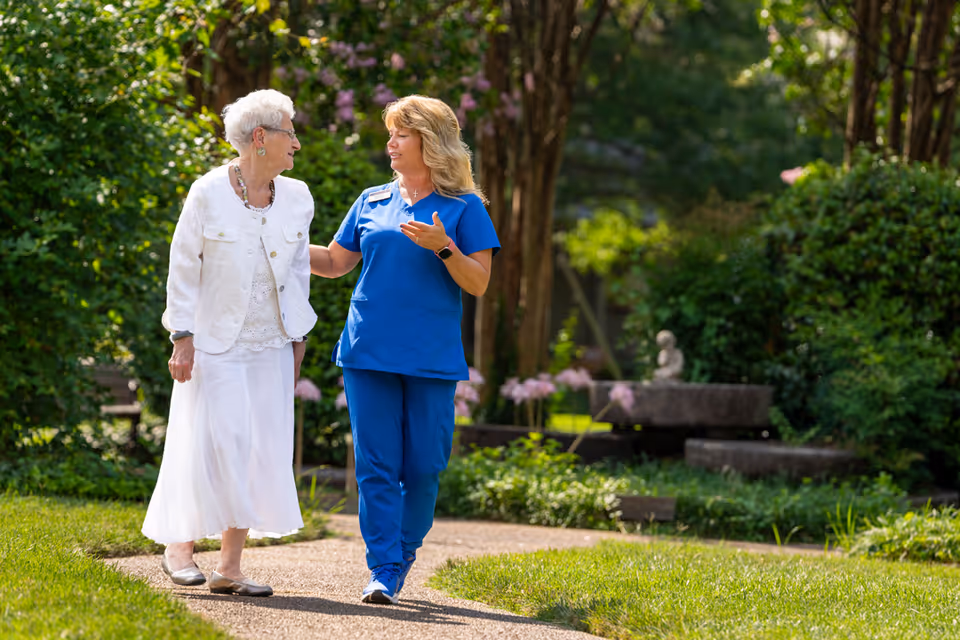 An elderly woman dressed in white walks along a garden path accompanied by a caregiver in blue scrubs. They are engaged in conversation and surrounded by lush greenery and flowering plants in a peaceful outdoor setting.