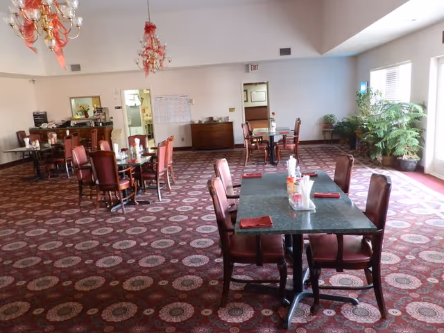 A spacious dining room with multiple tables and chairs arranged on a patterned carpet. The tables are set with napkins and condiments. There are large windows on the right side letting in natural light, and several potted plants near the windows. Chandeliers hang from the ceiling, and there is a sideboard with items on it against the far wall.