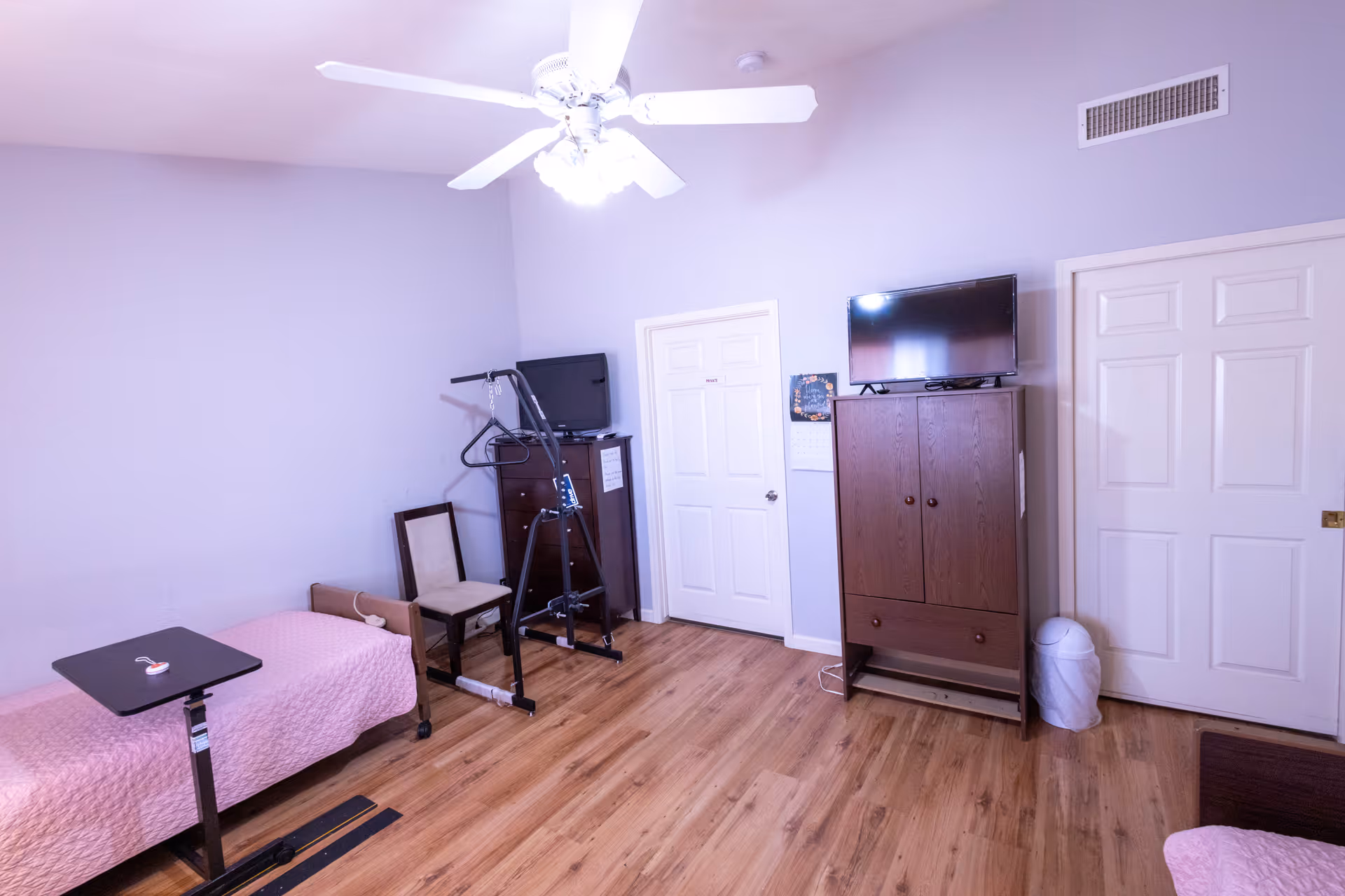 A senior living facility bedroom with a single bed covered in a pink quilt, a small adjustable table beside the bed, a wooden chair, a dark wooden dresser with a TV on top, and a wooden cabinet with another TV on top. The room has light purple walls, wooden flooring, a ceiling fan with lights, and two white doors. There is also a trash can near one of the doors.