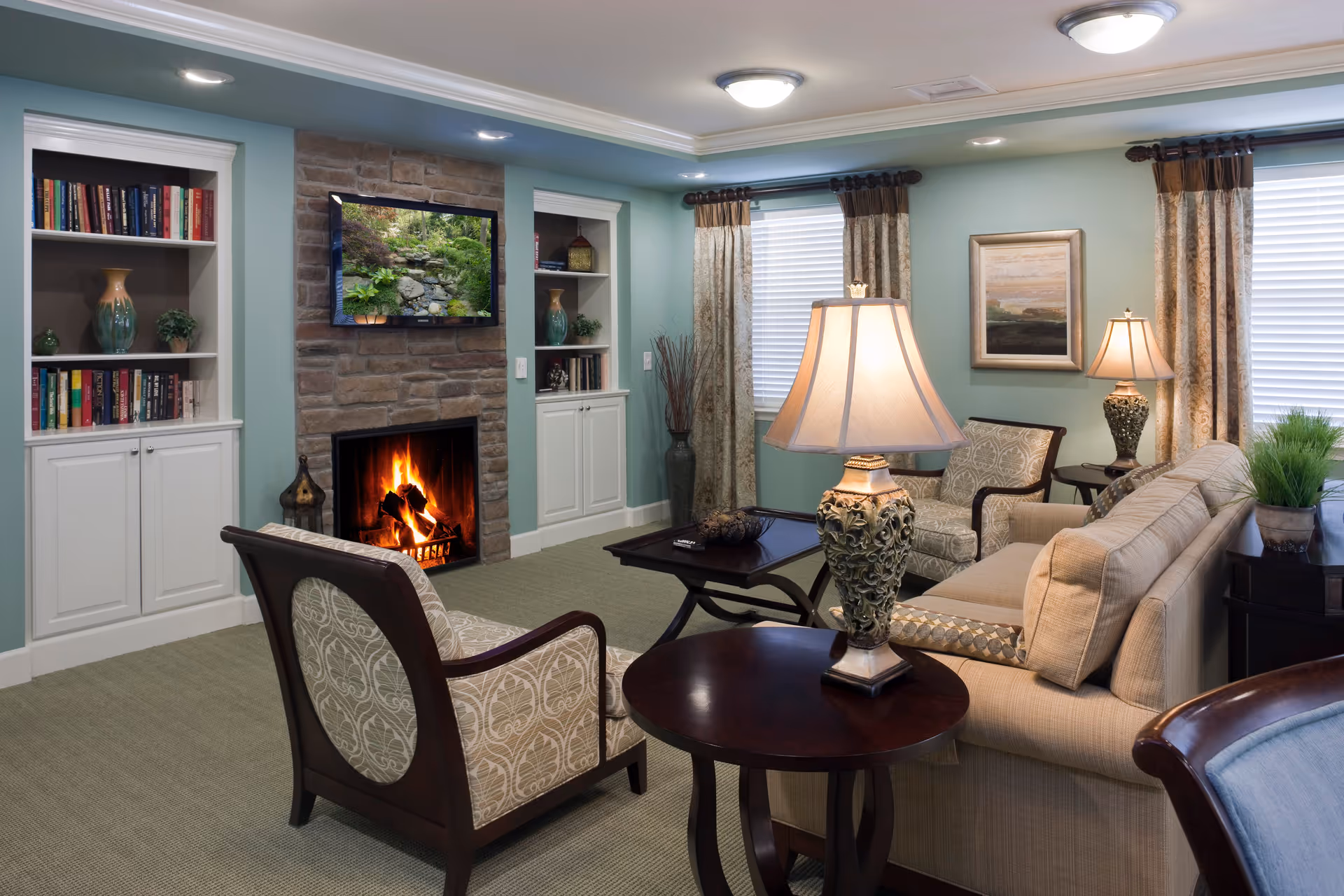 A cozy living room with a lit stone fireplace, a wall-mounted TV above it displaying a nature scene, built-in white bookshelves on either side, and comfortable seating including a beige sofa and patterned armchairs. The room has light blue walls, two windows with curtains, a dark wood coffee table, and two decorative table lamps.