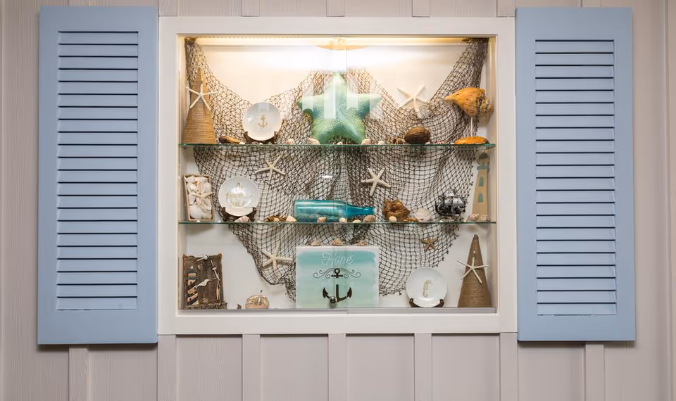 A decorative wall display with two light blue shutter panels on either side. Inside the display case are two glass shelves holding various nautical-themed items including starfish, seashells, a green star-shaped ornament, a small lighthouse model, a bottle, and plates with anchor and seahorse designs, all arranged against a backdrop of fishing net.