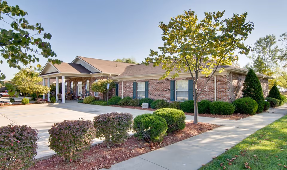 Exterior view of a single-story brick building with a covered entrance supported by white columns. The building is surrounded by neatly trimmed bushes, small trees, and a concrete sidewalk. The sky is clear and the scene is well-lit by daylight.
