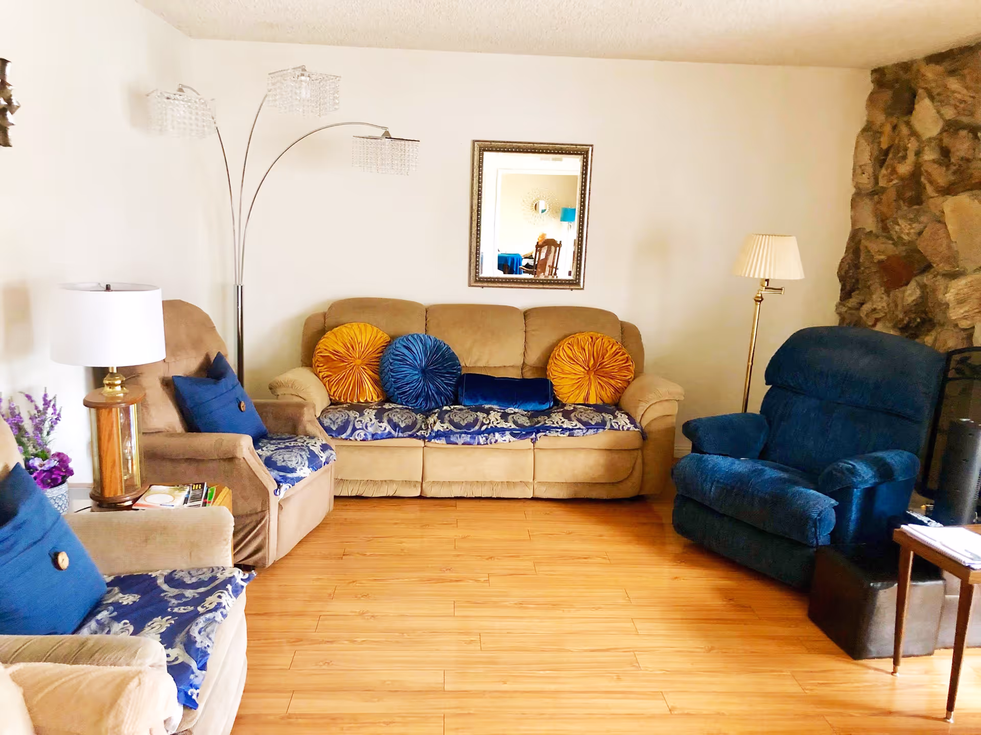 A cozy living room with a beige sofa adorned with blue and orange round pillows, two beige armchairs with blue cushions and patterned covers, a blue recliner chair, a wooden floor, a stone accent wall on the right, a floor lamp with three lights, a table lamp on a side table, and a mirror on the wall reflecting part of another room.